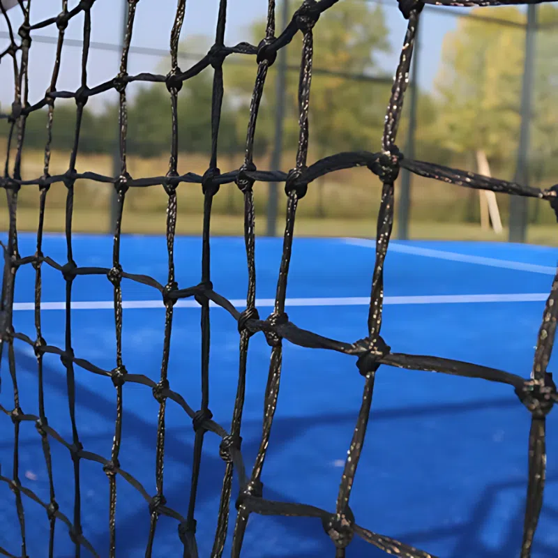 Una vista en primer plano de una red negra en una cancha de tenis al aire libre de color azul capta la atención. En el fondo, una pelota Red Paddle Uk Time Sport de 10 m x 0,92 m reposa sobre el césped entre árboles ligeramente borrosos y un cielo despejado, lo que sugiere que se aproxima un partido.