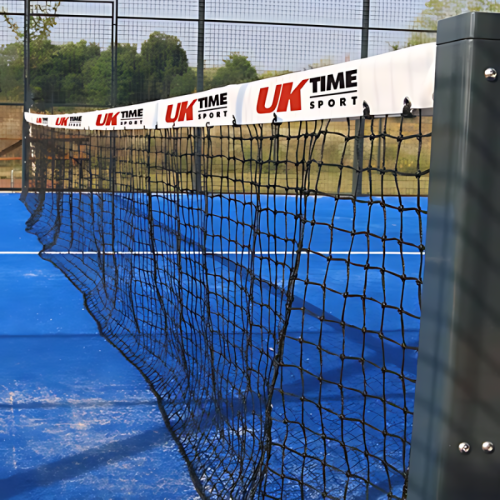 En un día despejado, una vista en primer plano captura la red de una cancha de tenis o pádel con su superficie azul visible. La red, marcada de forma destacada con la marca "UK TIME Sport" en la cinta blanca de la parte superior, mide 10 m x 0,92 m y se destaca claramente. Cerca, se coloca el equipo de Red Paddle, mientras que los árboles y las vallas enmarcan el fondo de la escena.