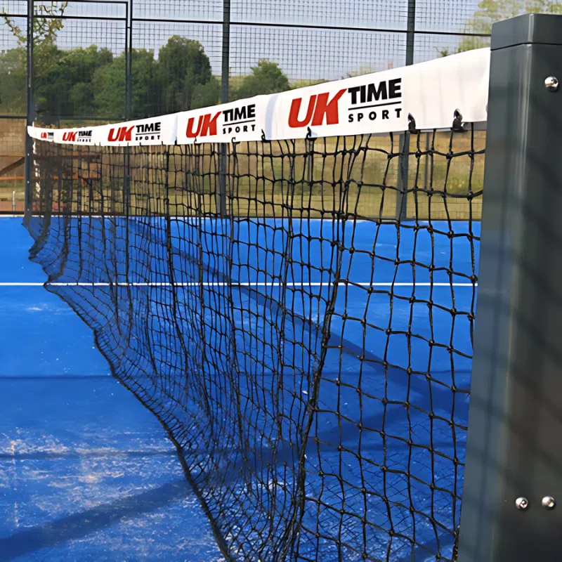 En un día despejado, una vista en primer plano captura la red de una cancha de tenis o pádel con su superficie azul visible. La red, marcada de forma destacada con la marca "UK TIME Sport" en la cinta blanca de la parte superior, mide 10 m x 0,92 m y se destaca claramente. Cerca, se coloca el equipo de Red Paddle, mientras que los árboles y las vallas enmarcan el fondo de la escena.