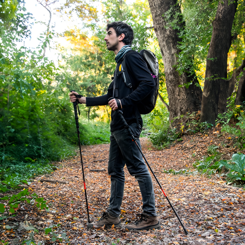 Un hombre con una mochila se encuentra de pie en un sendero cubierto de hojas en un bosque, sosteniendo Bastones de Trekking Plegables y Telescópicos Kano. Los árboles circundantes son frondosos y la luz del sol se filtra a través del follaje, creando una atmósfera pacífica.