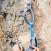 La mano del escalador agarra firmemente una pared rocosa irregular, con una cinta exprés Cinta Express Petzl Djinn Axess unida a un perno por encima. La cinta exprés azul asegura la cuerda a la pared rocosa.