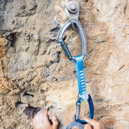 La mano del escalador agarra firmemente una pared rocosa irregular, con una cinta exprés Cinta Express Petzl Djinn Axess unida a un perno por encima. La cinta exprés azul asegura la cuerda a la pared rocosa.