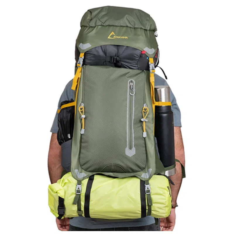 Alguien que lleva la Mochila Atakama Outdoor Torres del Paine 60+10L muestra su espacioso diseño verde para senderismo, con múltiples compartimentos, detalles en amarillo y equipo adjunto. La mochila contiene una botella de agua de metal y un saco de dormir enrollado. Están orientados hacia el otro lado para enfatizar el estilo distintivo de la mochila.