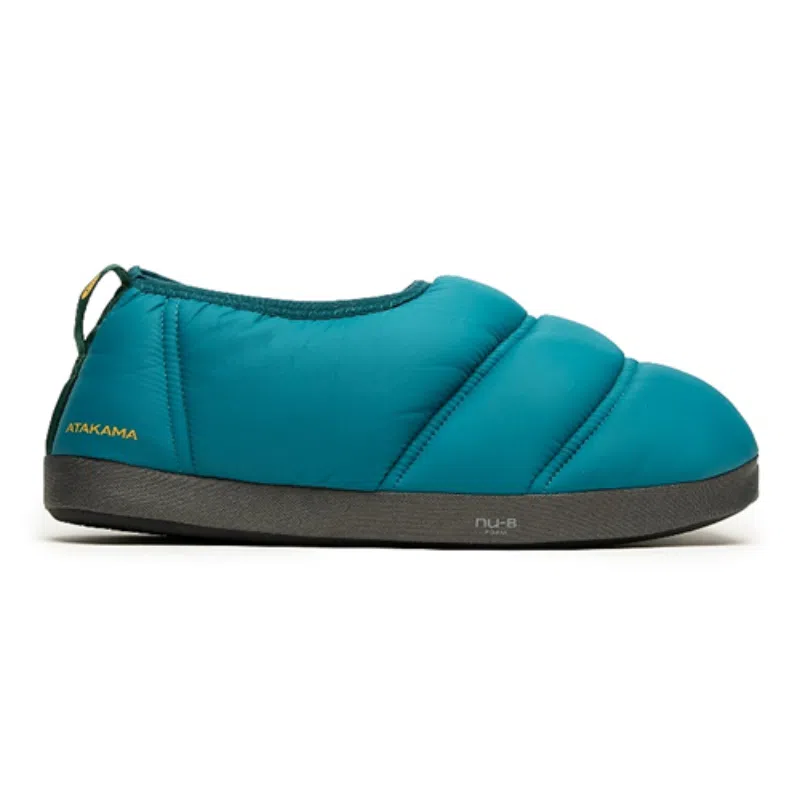Pantuflas de color verde azulado con diseño acolchado, suela negra y tirador. El nombre "Atakama" se muestra cerca del talón en el costado de la pantufla. Estas Pantuflas Térmicas Atakama Outdoor Futaleufú Nu-B Foam acogedoras y aislantes son adecuadas para uso en interiores, pero lo suficientemente resistentes para viajes rápidos al aire libre a Futaleufú.