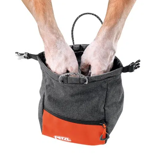 Las manos se introducen en una Bolsa Petzl para Magnesio Sakab, que muestra su diseño con un bolsillo naranja y un cuerpo negro adornado con el logotipo "Petzl". Las manos cubiertas de tiza insinúan la participación en actividades de escalada o gimnasia.