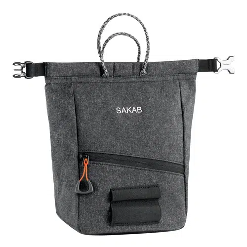 Bolsa portátil de tela gris con la marca "SAKAB" impresa en el frente, similar a la Bolsa Petzl para Magnesio Sakab. Incluye un pequeño bolsillo frontal con cremallera, dos asas tipo lazo y dos hebillas ajustables de liberación lateral en ambos lados. Además, hay una pequeña correa elástica ubicada debajo del bolsillo con cremallera.