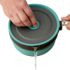 Una persona vierte agua de una olla plegable Sea To Summit Frontier de 1,3 l, que es una tetera de silicona plegable de color verde azulado y gris con un pico negro. Tiene un asa de metal y una tapa gris, y la persona sostiene la tapa firmemente con una mano mientras vierte.
