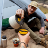 Un hombre se sienta junto a una tienda de campaña sobre una superficie arenosa, vertiendo una bebida caliente de su tetera Sea To Summit Frontier en una taza a juego del Set de Cocina de 3 piezas. A su lado se ven otros elementos para acampar al aire libre, como una cantimplora y otra taza, con un bosque y un cuerpo de agua de fondo.