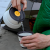 Una persona usa una tetera plegable del Set de Cocina 3pc Sea To Summit Frontier para verter agua caliente en una taza. De pie cerca de su tienda de campaña, bien abrigados para protegerse del frío, saborean la serena calidez de la aventura al aire libre.