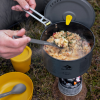 Mientras una persona cocina un risotto en una estufa portátil para acampar al aire libre, una olla gris hierve a fuego lento mientras revuelven hábilmente con una cuchara Cuchara Sea To Summit Mango Largo Frontier, manteniendo la tapa firme. Cerca, sobre el césped, descansa un pequeño tazón amarillo y una taza, que completan esta tranquila escena de cena en la frontera.