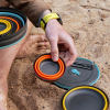 Mientras alguien se relaja en la arena, coloca cuencos de camping coloridos y apilables, como la Taza Plegable de Sea To Summit Frontier de 400 ml. Los cuencos tienen tonos naranja, amarillo y turquesa y están fabricados con material plegable. Un reloj adorna su muñeca mientras se pueden ver sus pies descalzos en esta configuración de aventura.