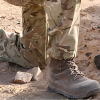 Una persona que viste pantalones de camuflaje militar y botas de combate marrones se arrodilla sobre una superficie rocosa y arenosa al aire libre, mostrando la robusta durabilidad de la bota ligera táctica Calcetín Teko 2.0.