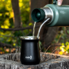 Un elegante Kano Mate Térmico Acero Inoxidable vierte líquido en una taza negra etiquetada "KANO" sobre un tocón de árbol, sobre un fondo de vegetación borrosa y luz solar que sugiere un ambiente tranquilo al aire libre.