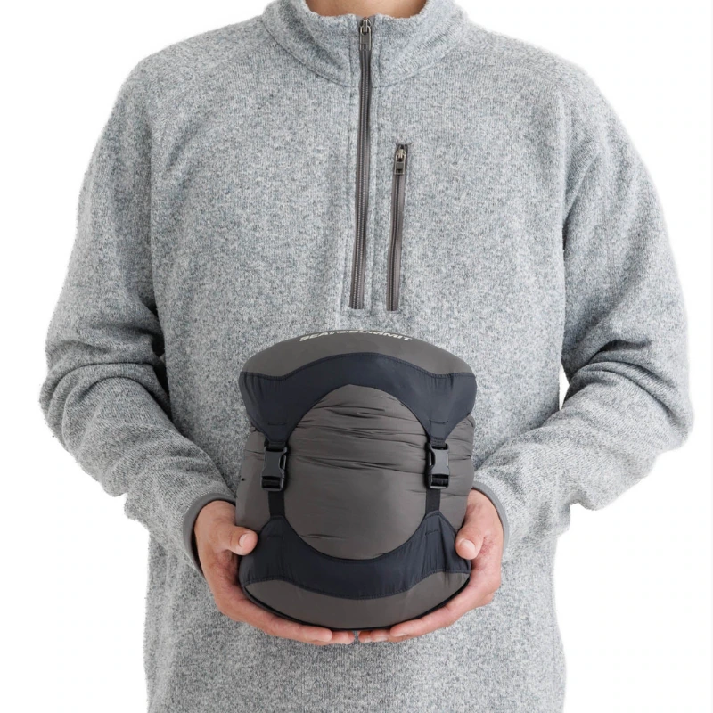 Una persona con una chaqueta de polar gris sostiene un Saco de Dormir Pluma Sea to Summit Spark Pro enrollado de forma compacta, asegurado con correas negras y que presenta un elegante diseño gris con detalles en negro.