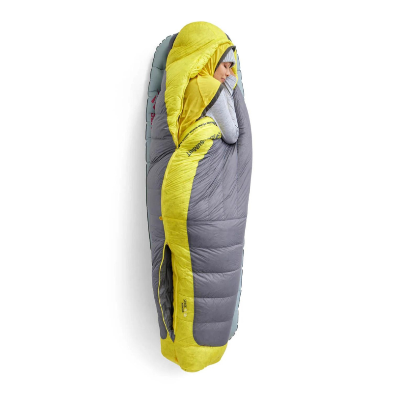 Una persona se encuentra de pie, cómodamente envuelta en un saco de dormir Pluma Sea To Summit Spark Women's -9°C. El diseño amarillo y gris, con la capucha sobre la cabeza y los brazos metidos dentro, crea una apariencia de capullo contra el fondo blanco.