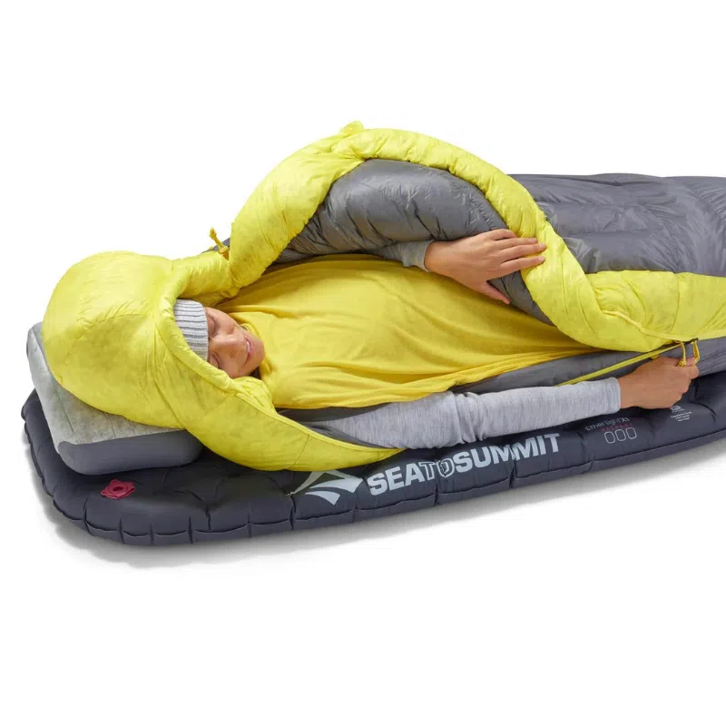 Una persona se encuentra acurrucada dentro de un saco de dormir Pluma Sea To Summit Spark Women's -9°C sobre una colchoneta aislante. El saco de dormir está parcialmente abierto, lo que deja al descubierto el rostro y las manos de la persona mientras descansa cómodamente con la marca Sea to Summit claramente visible en la colchoneta.