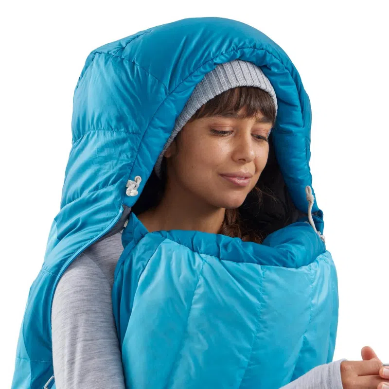 Una persona que lleva un gorro gris y un suéter gris claro está cómodamente envuelta en un saco de dormir azul Sea To Summit Trek -9C con capucha. El saco de dormir se ajusta cómodamente alrededor de su rostro, lo que sugiere calidez y comodidad.