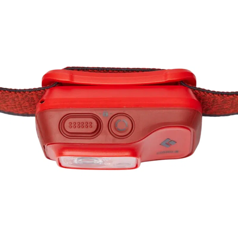 Una linterna frontal roja Black Diamond con una correa de tela y varios controles en el lateral. Esta linterna frontal recargable cuenta con un botón de encendido prominente y un interruptor texturizado, probablemente para ajustar el brillo o los ajustes de modo. La parte frontal tiene una lente transparente para el LED.