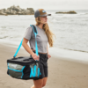 Una persona que viste una camiseta gris, pantalones cortos negros y una gorra camina por la playa con una hielera plegable Tyndall Soft Cooler Plegable Kano de 45 litros colgada del hombro. El océano y las rocas forman un fondo sereno bajo el cielo nublado.