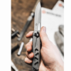 Una mano sostiene una Navaja Boker Plus Specialist Half-Tool 09BO831 abierta, mostrando su elegante diseño metálico, con un fondo borroso de herramientas y papeles sobre una superficie de madera que realza su atractivo.