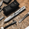 La herramienta Navaja Boker Plus Specialist Half-Tool 09BO831, con mango metálico plateado, reposa sobre una superficie de madera rústica. Cerca se encuentran sus accesorios: una bolsa negra, una lima y un tornillo de sujeción.