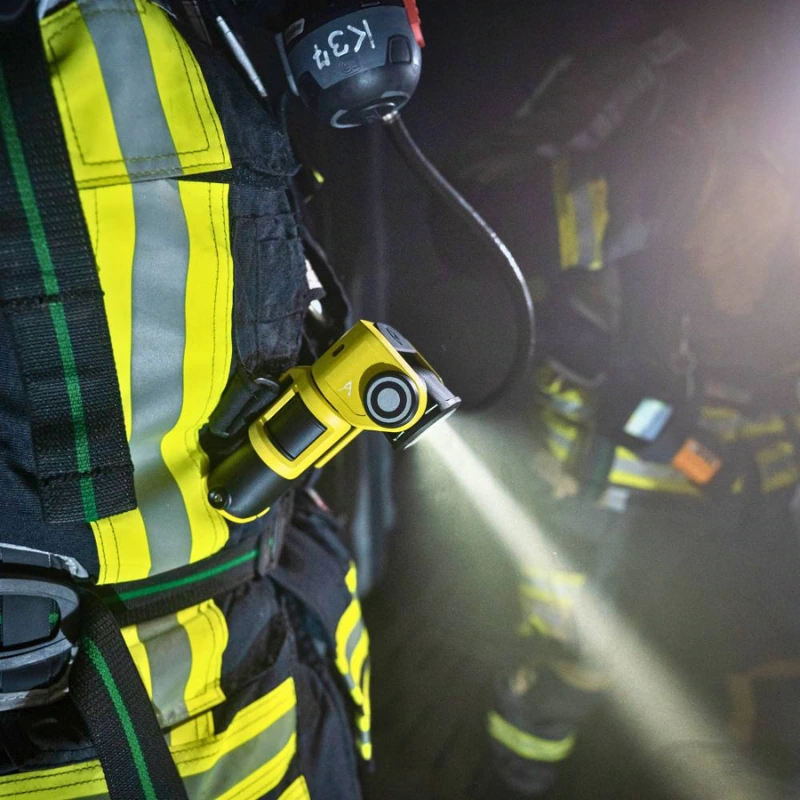 En el equipamiento de un bombero destaca la Linterna Recargable Led Lenser EXC6R Atex 300lm, una linterna de color amarillo brillante que garantiza la visibilidad. Al fondo, se ve parcialmente a otro bombero con ropa de protección con bandas reflectantes.