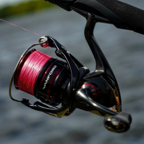 Primer plano de un carrete de pesca Carrete Shimano Vanford 1000F de 170 m/0,18 mm negro y rojo con hilo rosa, sobre un fondo de agua desenfocado. El nombre de la marca se ve claramente en el carrete.