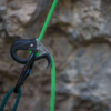 Un primer plano de un dispositivo de seguridad Black Diamond ATC-Pilot Descensor y un mosquetón unidos a una cuerda de escalada verde resalta el equipo de escalada contra un fondo rocoso borroso, capturando perfectamente la esencia de un entorno de escalada en roca aventurero.