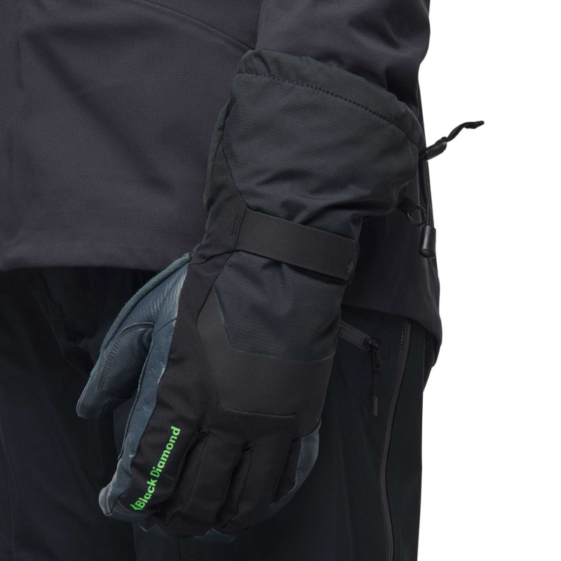 Una persona lleva una chaqueta oscura con cremallera y un par de guantes Black Diamond Crag Gloves (copia), que presentan un texto verde y una combinación de materiales oscuros y claros. Los guantes están equipados con un puño ajustable para garantizar un ajuste perfecto.