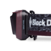 Primer plano de una linterna frontal que muestra el texto "DUAL FUEL" en el lateral. La correa muestra parte de un logotipo. Este dispositivo, identificado como Linterna de Cabeza Black Diamond Astro 300, es principalmente de color negro y granate.