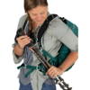Una persona vestida con una camisa gris y pantalones azules ajusta unos bastones de trekking conectados a su mochila verde Mochila Osprey Talon 22 (copia) mientras mira los bastones contra un fondo blanco liso.