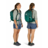 Dos vistas de una persona que lleva una mochila Osprey Talon 22 (copia) verde azulado: vista frontal y lateral. Lleva una camiseta gris claro, pantalones cortos estampados azules y zapatos gris oscuro. La mochila está diseñada con varias correas para brindar soporte.