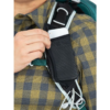 Una persona que viste una camisa a cuadros desliza un teléfono inteligente en una pequeña bolsa negra sujeta a la correa de su mochila Mochila Osprey Talon 22 (copia), con auriculares blancos envueltos alrededor de la correa.