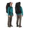 Un hombre muestra la Mochila Osprey Aether 65L mientras viste una camisa a cuadros verde, pantalones grises, zapatos de senderismo y una gorra negra con una parte superior roja. Está fotografiado tanto de frente como de perfil sobre un fondo blanco liso.