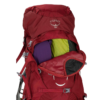 Mochila para mujer Osprey Ariel 65 roja con un compartimento superior abierto que muestra los artículos guardados, incluida la tela verde y violeta. La mochila está equipada con múltiples correas y una hebilla, lo que la hace ideal para practicar senderismo o viajar.