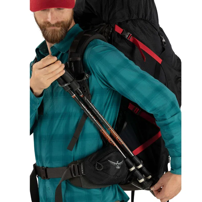 Una persona con una camisa a cuadros azul y una gorra roja lleva una mochila Osprey Aether Plus de 85 litros con correas y compartimentos visibles. Lleva bastones de trekking en la mano, lleva un cinturón con una pequeña bolsa adjunta y ajusta la correa de la mochila.