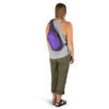 Una persona con cabello largo se encuentra de espaldas a la cámara, vistiendo una camiseta gris sin mangas, pantalones verde oliva y sandalias negras. Lleva una Mochila Bandolera Osprey Daylite Sling morada, lo que le da un toque de aventura a su look.