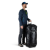 Una persona que viste una chaqueta azul, pantalones oscuros y una gorra sostiene el asa de una bolsa de lona con ruedas Osprey Transporter 60L, mostrando con orgullo el elegante logotipo en su gran superficie negra.