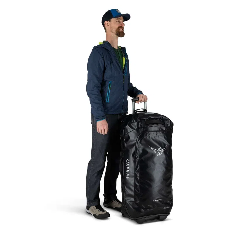 Una persona que viste una chaqueta azul, pantalones oscuros y una gorra sostiene el asa de una bolsa de lona con ruedas Osprey Transporter 60L, mostrando con orgullo el elegante logotipo en su gran superficie negra.