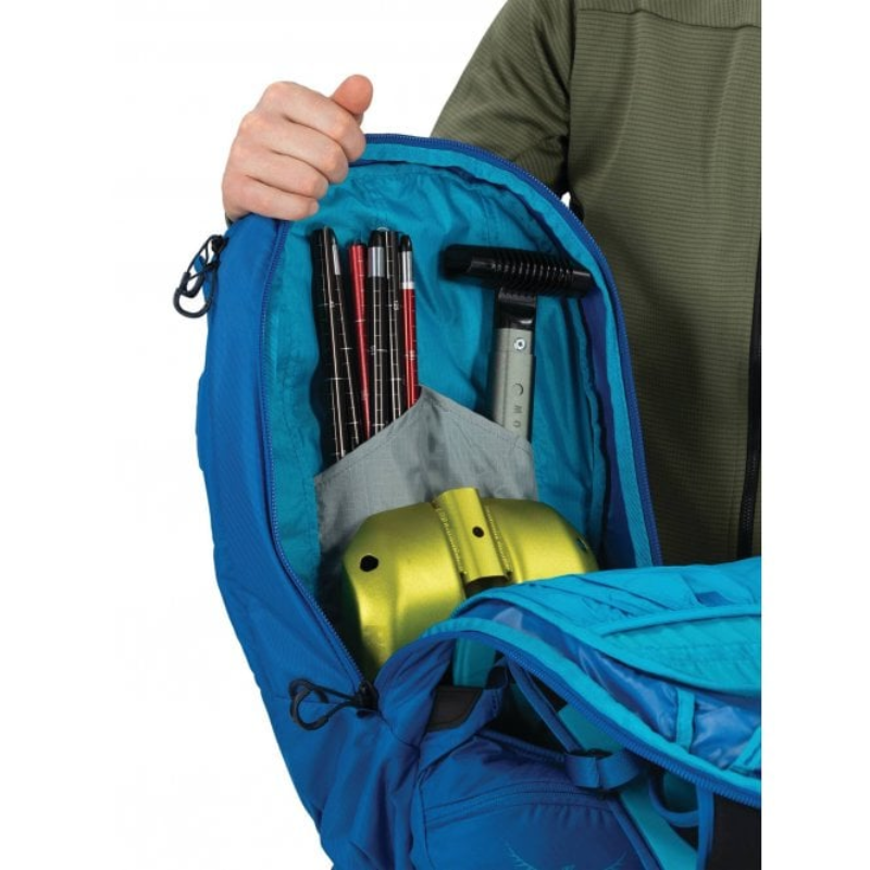Una persona abre la Mochila para Nieve Osprey Kamber 20 de color azul y descubre que su contenido está bien organizado. En el interior hay un juego de bastones, una herramienta negra y un objeto de color amarillo brillante. La persona lleva una chaqueta verde y solo se le ven la mano y parte del brazo.