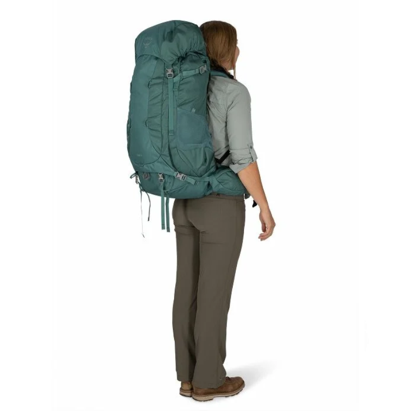Una persona que lleva una mochila de viaje Osprey Viva 65 Mujer de color verde azulado de gran tamaño está de espaldas. Lleva una camisa de manga larga de color gris claro, pantalones de color verde oliva y zapatos marrones. La mochila está completamente llena y su tamaño supera con creces la cabeza de la persona.