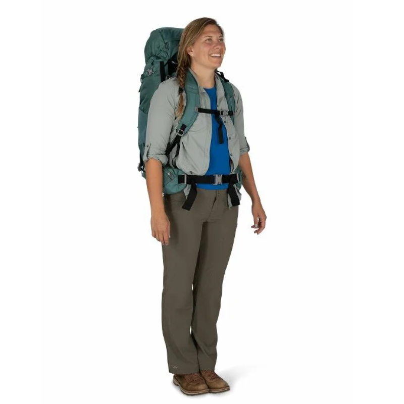 Una persona vestida con ropa de calle sostiene una mochila grande Osprey Viva 65 Mujer. Sonríe y mira hacia un lado. La mochila es de color verde azulado, mientras que la persona lleva una camiseta gris claro, pantalones marrones y zapatos marrones. El fondo es blanco liso.
