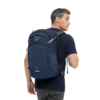 Una persona con cabello corto y gris lleva una mochila de montaña Osprey Mutant 38 (copia) azul marino. Está vestida con una camiseta azul oscuro y pantalones grises, y mira por encima del hombro hacia la derecha contra un fondo blanco liso.