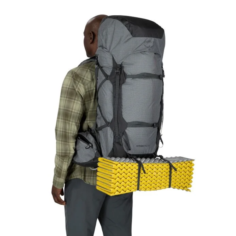 El individuo, que lleva una mochila Osprey Aether Pro de 75 litros, lleva una camisa a cuadros y pantalones grises. Con una colchoneta amarilla doblada sujeta a la parte inferior de la gran mochila gris, está de espaldas a la cámara, listo para la aventura.