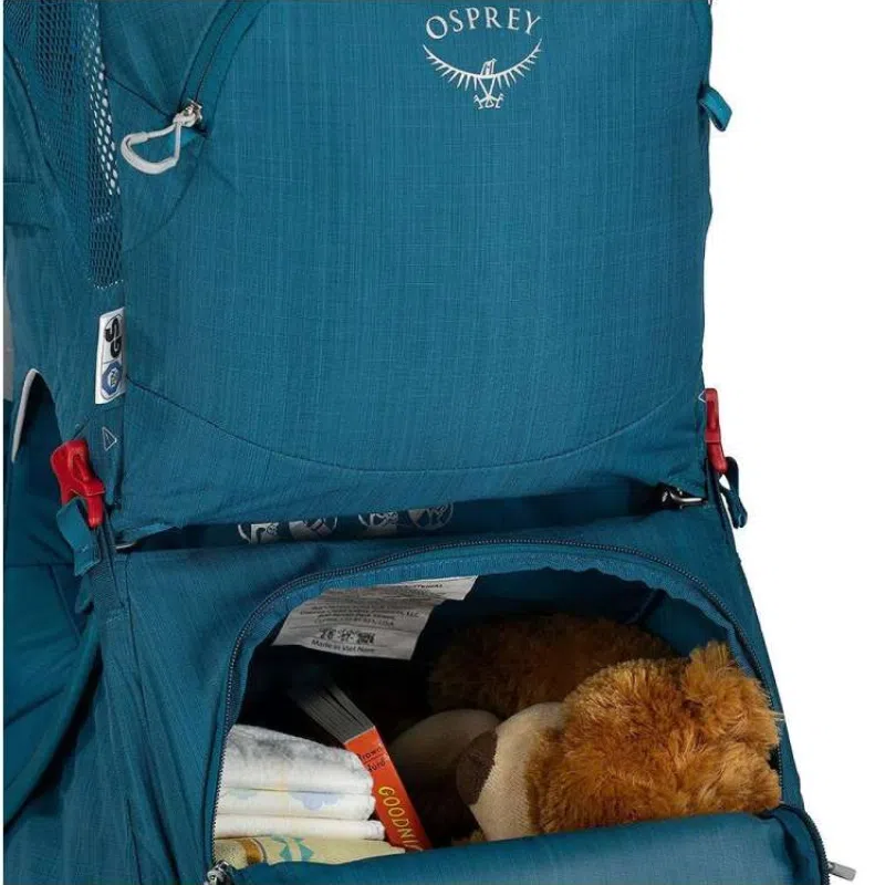 Una mochila portabebé azul Osprey Poco SLT con un compartimento abierto revela un osito de peluche marrón, una manta de bebé y un libro infantil titulado "Buenas noches".