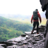 Un excursionista con una chaqueta roja, protegido por su Funda Impermeable Osprey Ultralight Raincover (copia), avanza por un sendero rocoso a lo largo de un borde escarpado, envuelto por la niebla y la lluvia. Se pueden ver exuberantes colinas verdes al fondo.