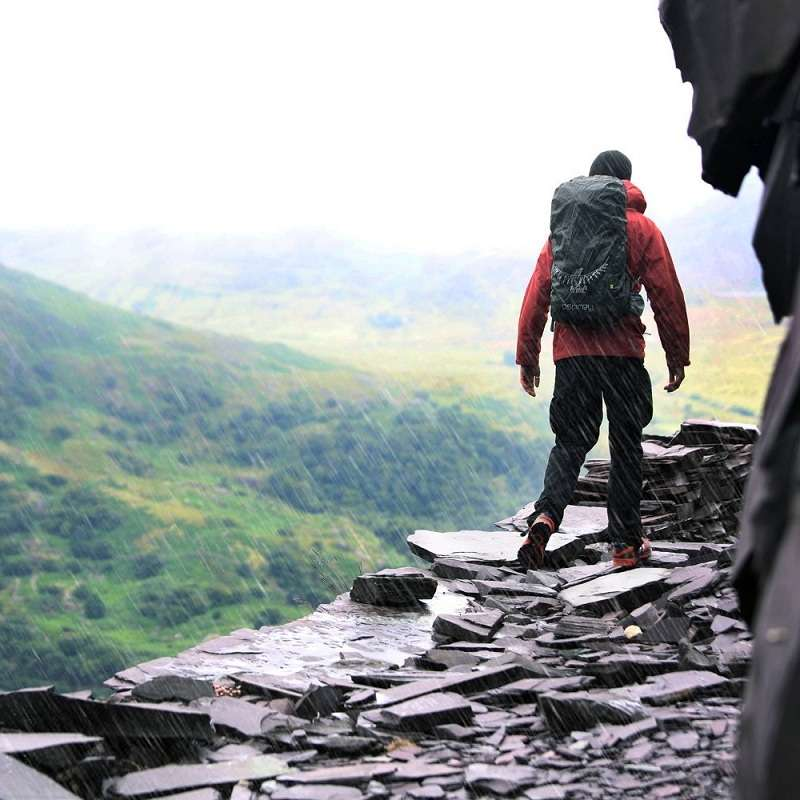 Un excursionista con una chaqueta roja, protegido por su Funda Impermeable Osprey Ultralight Raincover (copia), avanza por un sendero rocoso a lo largo de un borde escarpado, envuelto por la niebla y la lluvia. Se pueden ver exuberantes colinas verdes al fondo.