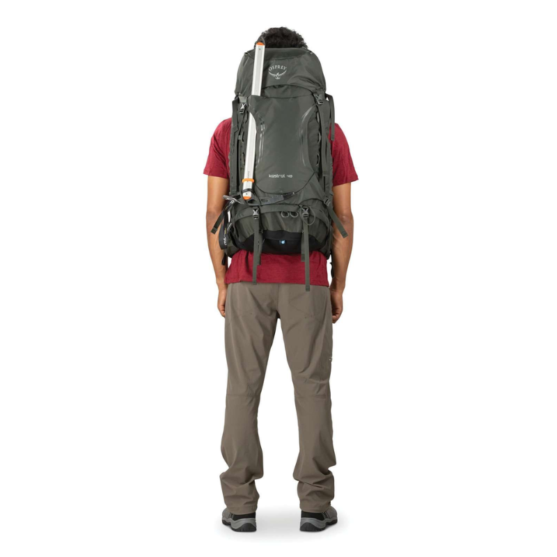 Una persona, vista desde atrás, lleva equipo para actividades al aire libre y una mochila de senderismo Osprey Kestrel 38 (copia) de gran tamaño de color gris con un piolet adjunto. Está vestida con una camiseta roja y pantalones grises.