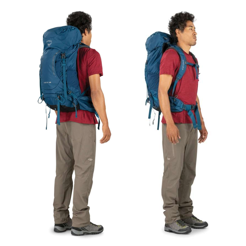 Una persona con una camiseta roja y un pantalón gris sostiene la Mochila de Viaje Osprey Farpoint 55 (copia) azul de gran tamaño desde dos ángulos, uno mirando hacia la izquierda y el otro hacia delante. La mochila está equipada con múltiples correas, lo que la hace ideal para practicar senderismo o para aventuras al aire libre.