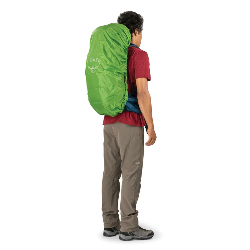 Una persona está de espaldas a la cámara, vestida con una camisa roja, pantalones beige y un par de zapatos para caminar. Lleva una mochila verde grande con una funda para la lluvia en la espalda, una opción ideal para el espíritu aventurero, similar a la confiable Mochila de Viaje Osprey Farpoint 55 (copia).
